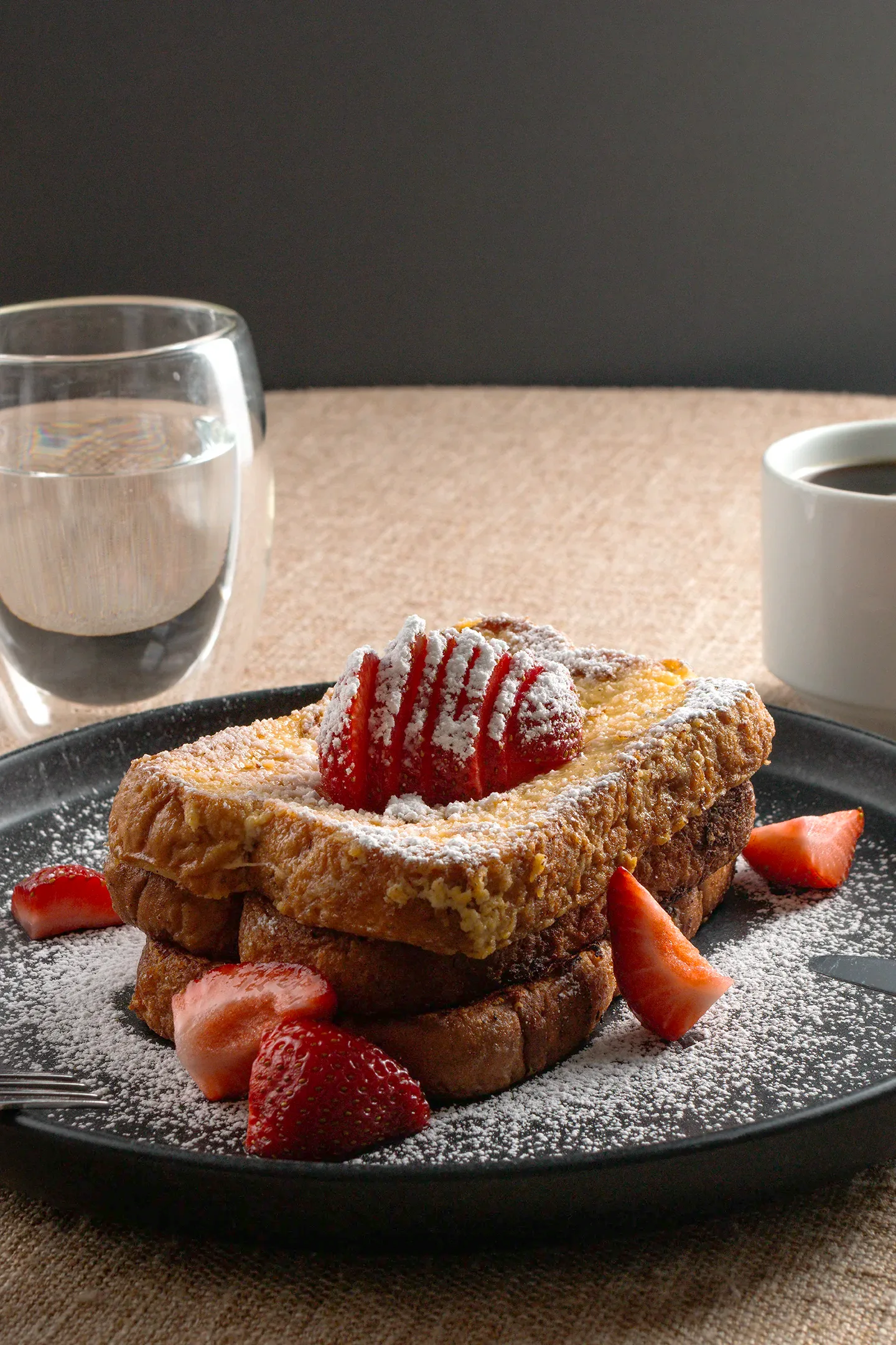 Delicioso pan francés con fresas y azúcar glas, servido en un elegante plato negro con café y agua, capturado por fotógrafo gastronómico en Chihuahua.