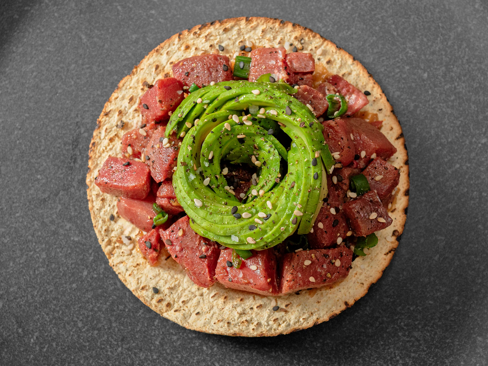 Fotografía cenital de una tostada con atún marinado en cubos y una espiral de aguacate en el centro, espolvoreada con semillas de sésamo y pimienta negra, sobre un plato oscuro.