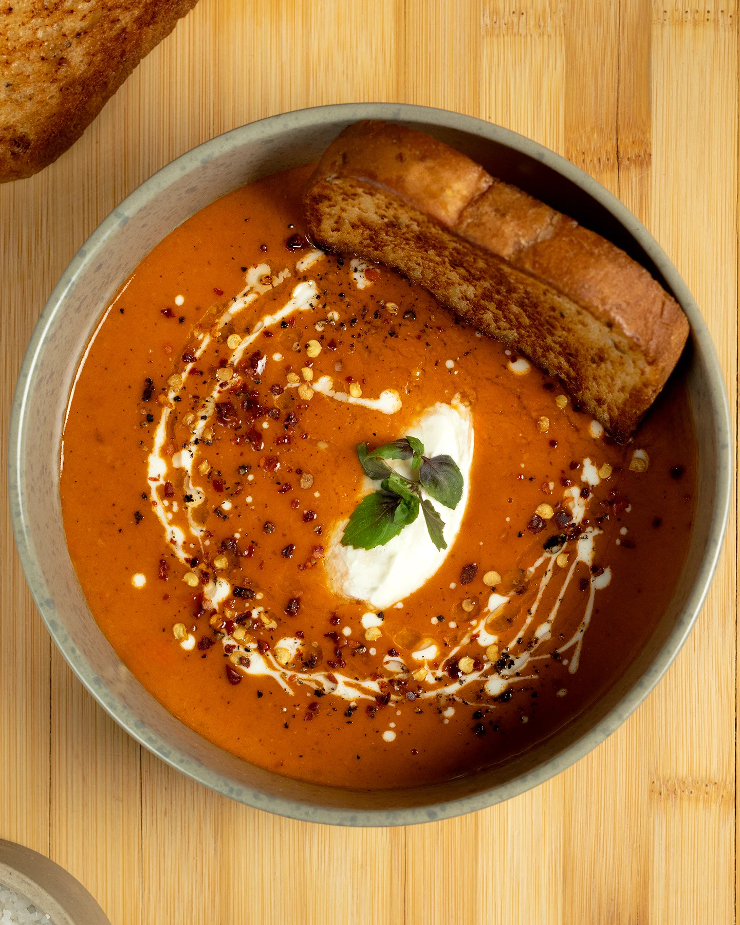 Sopa cremosa de tomate servida en bowl gris, decorada con crema, hojuelas de chile, pimienta negra y una ramita de microgreens, acompañada de pan tostado sobre fondo de madera clara.