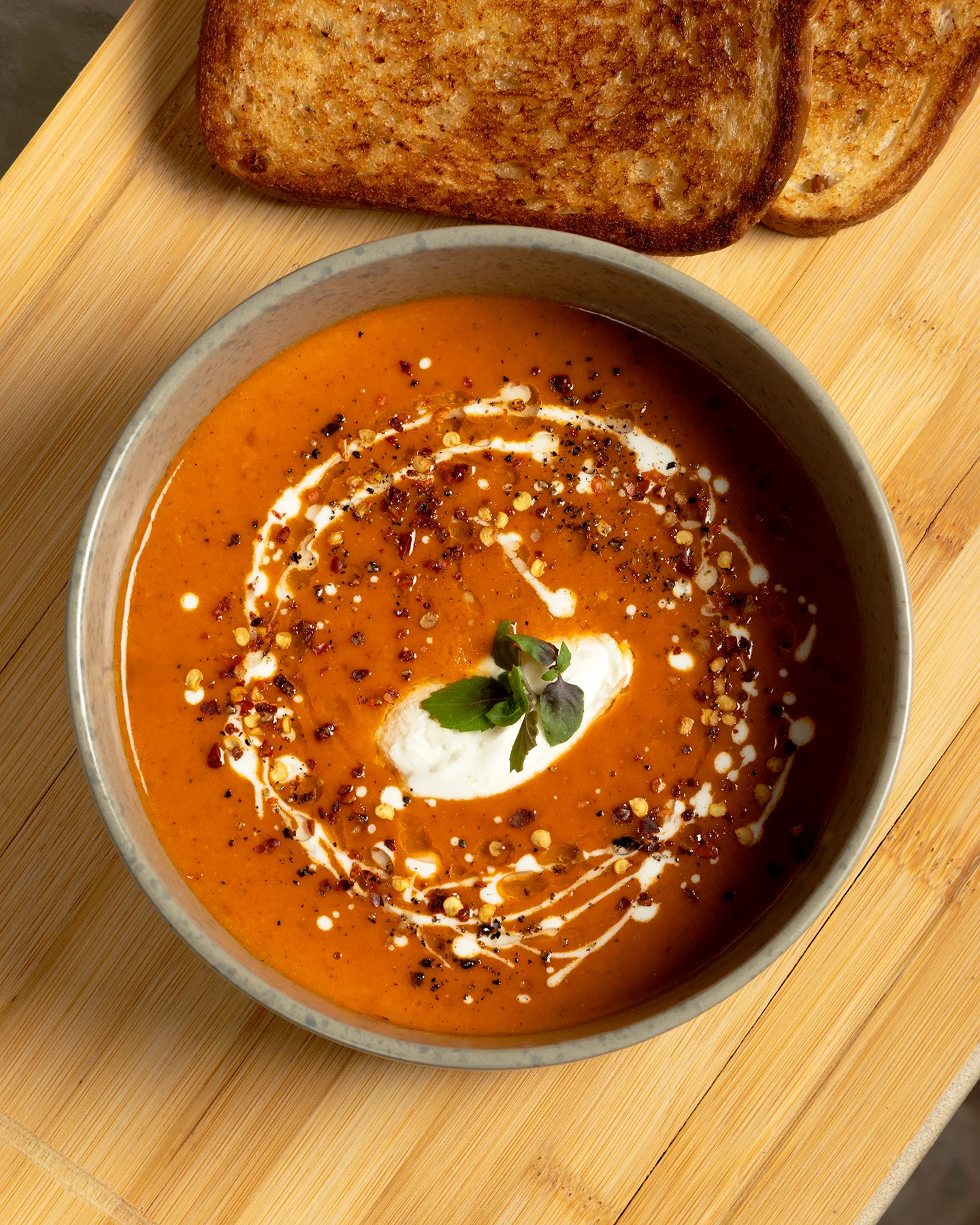 Sopa cremosa de tomate servida en bowl gris, decorada con crema, hojuelas de chile, pimienta negra y una ramita de microgreens, acompañada de pan tostado sobre fondo de madera clara.