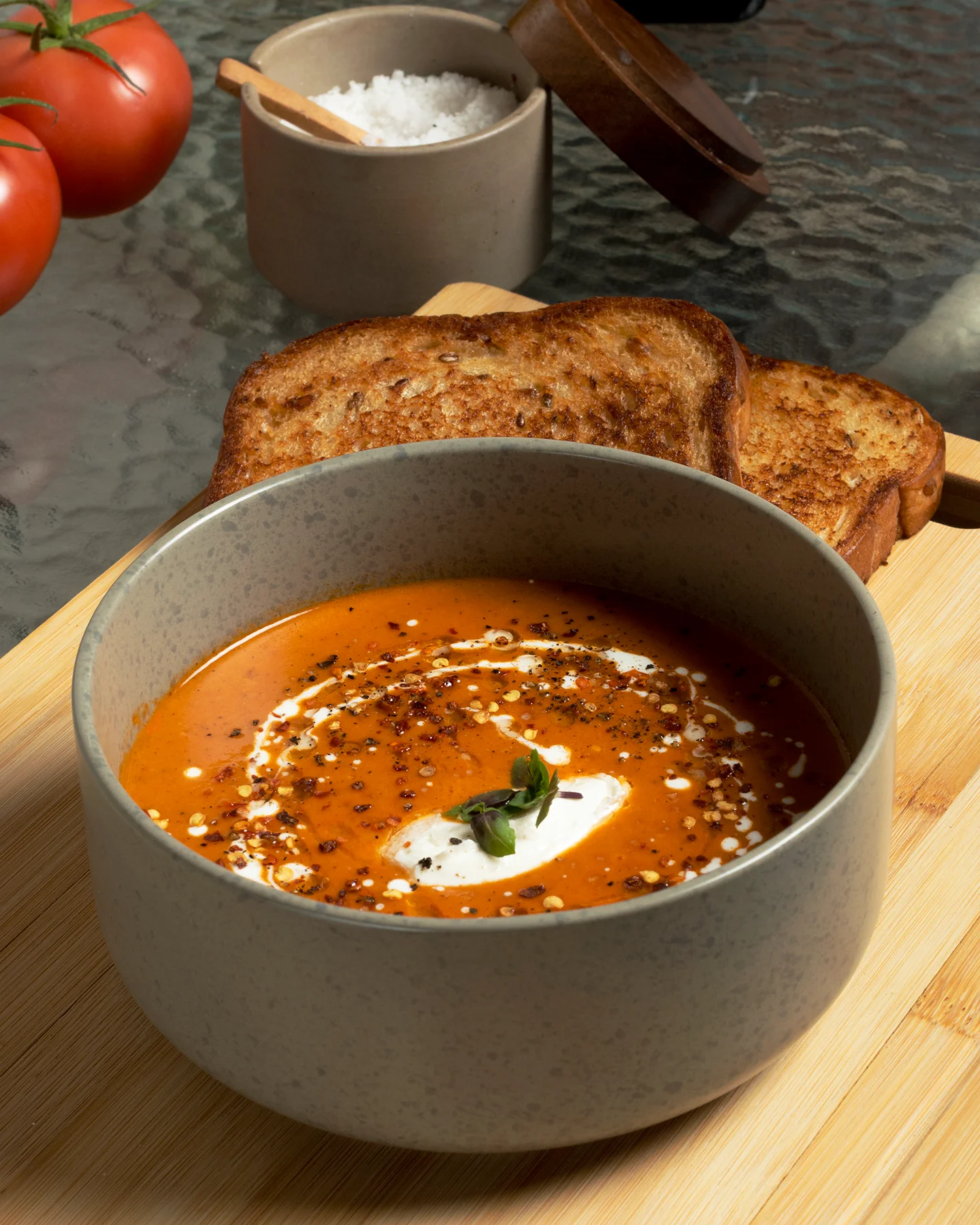 Fotografía gastronómica de una sopa de tomate servida en bowl gris, decorada con crema, hojuelas de chile, pimienta negra.