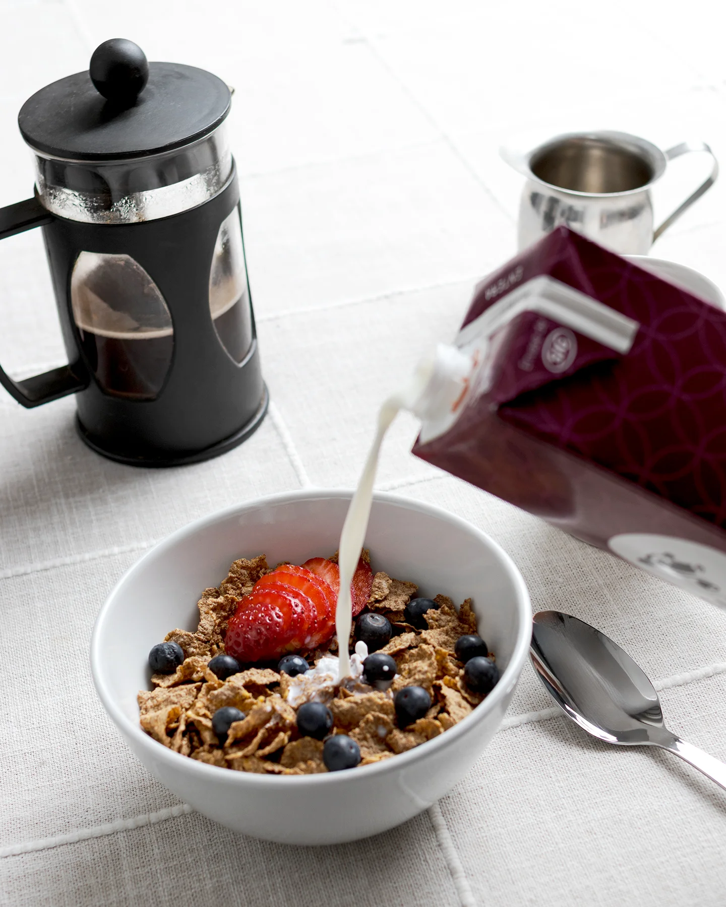 Tazón con cereal y frutas al que se vierte leche desde un envase, acompañado de prensa francesa y cuchara sobre mantel claro.