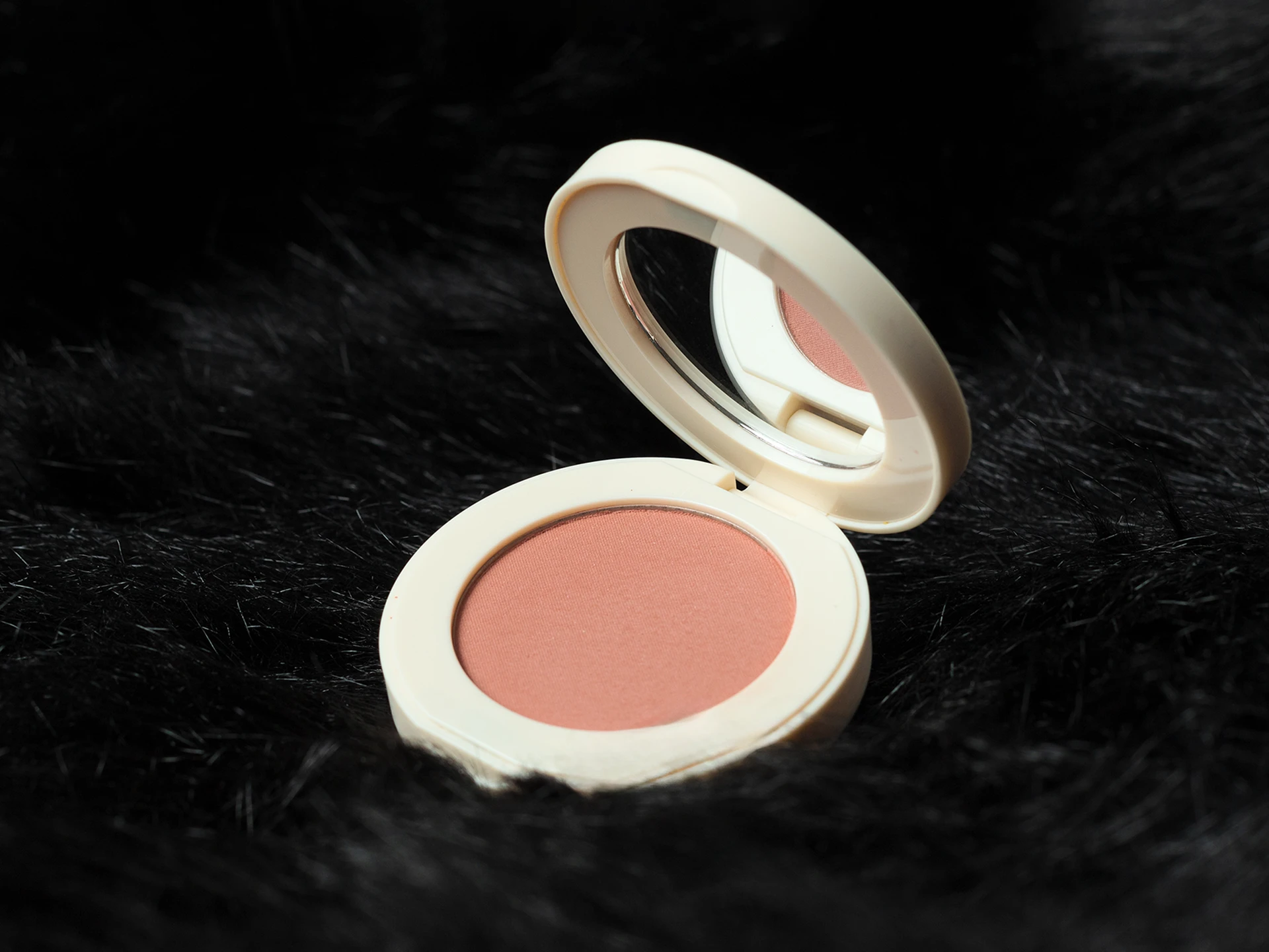 Rubor compacto en estuche beige con espejo abierto, tono rosa suave, sobre fondo de tela negra.