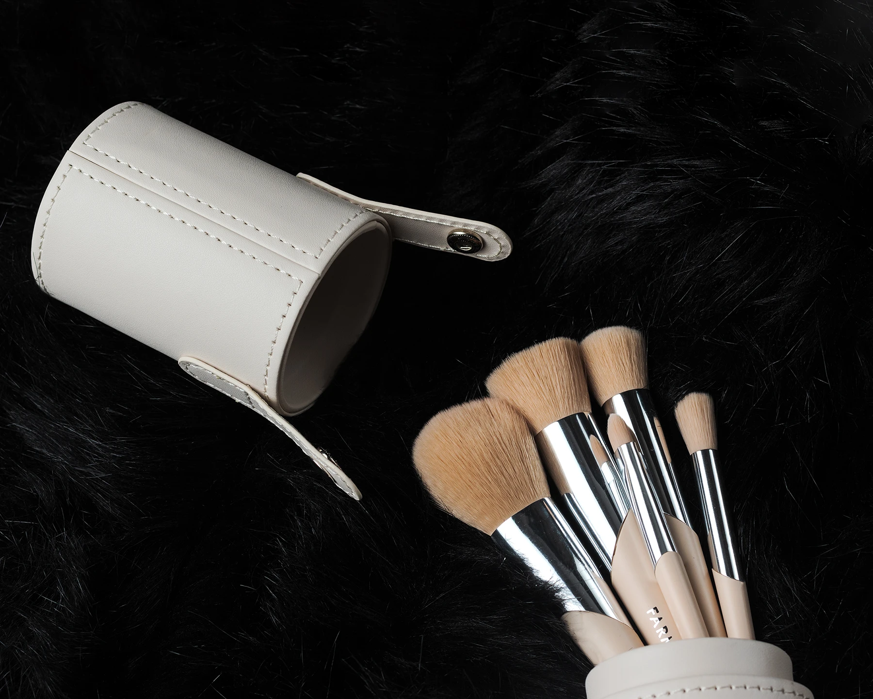 Set de brochas de maquillaje beige con estuche cilíndrico sobre fondo negro aterciopelado, fotografía de producto profesional