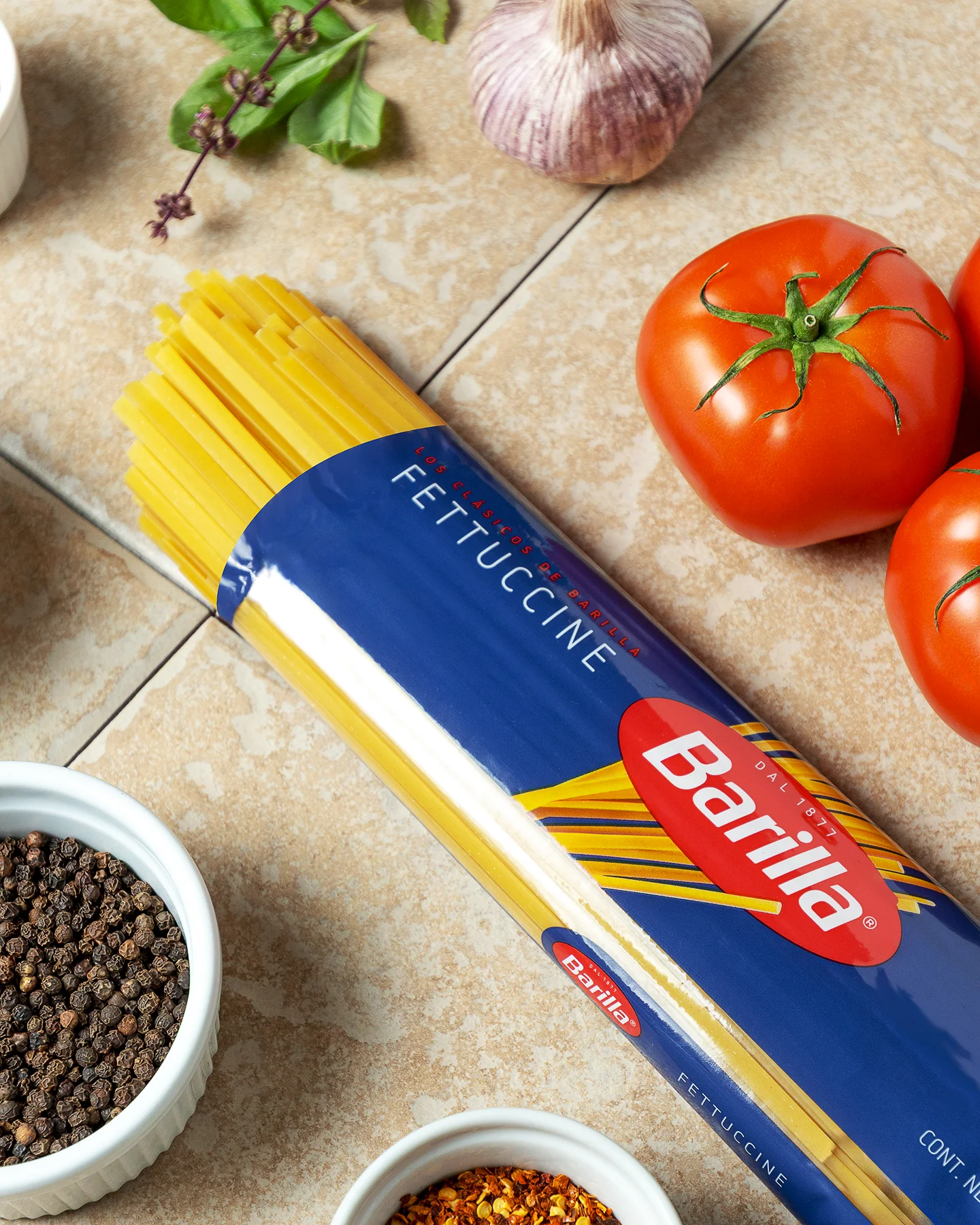 Fotografía publicitaria de un paquete de pasta Barilla Fettuccine rodeado de ingredientes: tomates, ajo, albahaca y especias en tazones blancos.