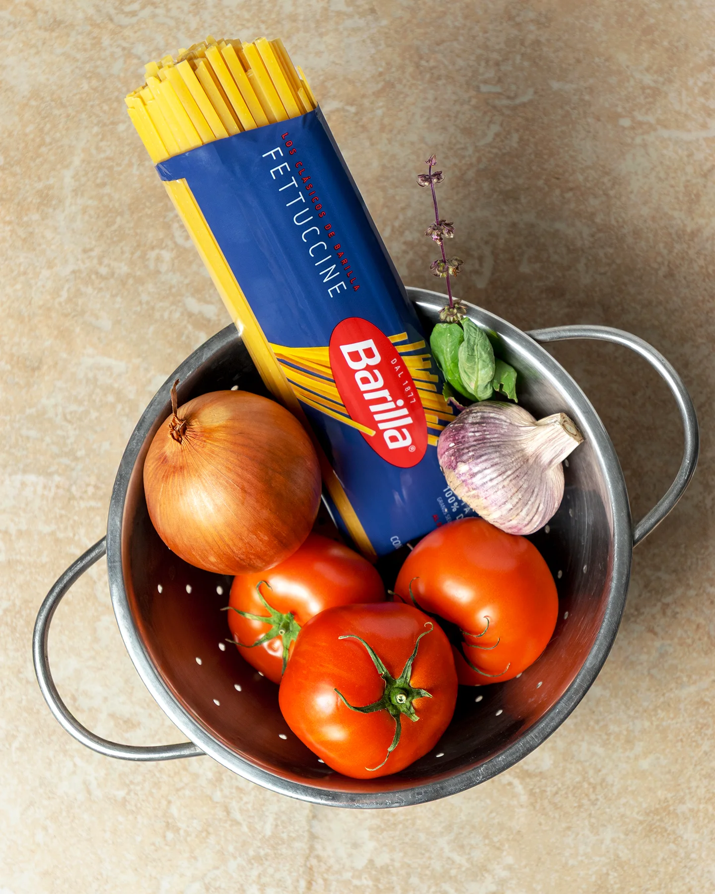 Fotografía en plano cenital de un colador de metal lleno de tomates, cebolla, ajo y albahaca, con un paquete de Barilla Fettuccine inclinado.