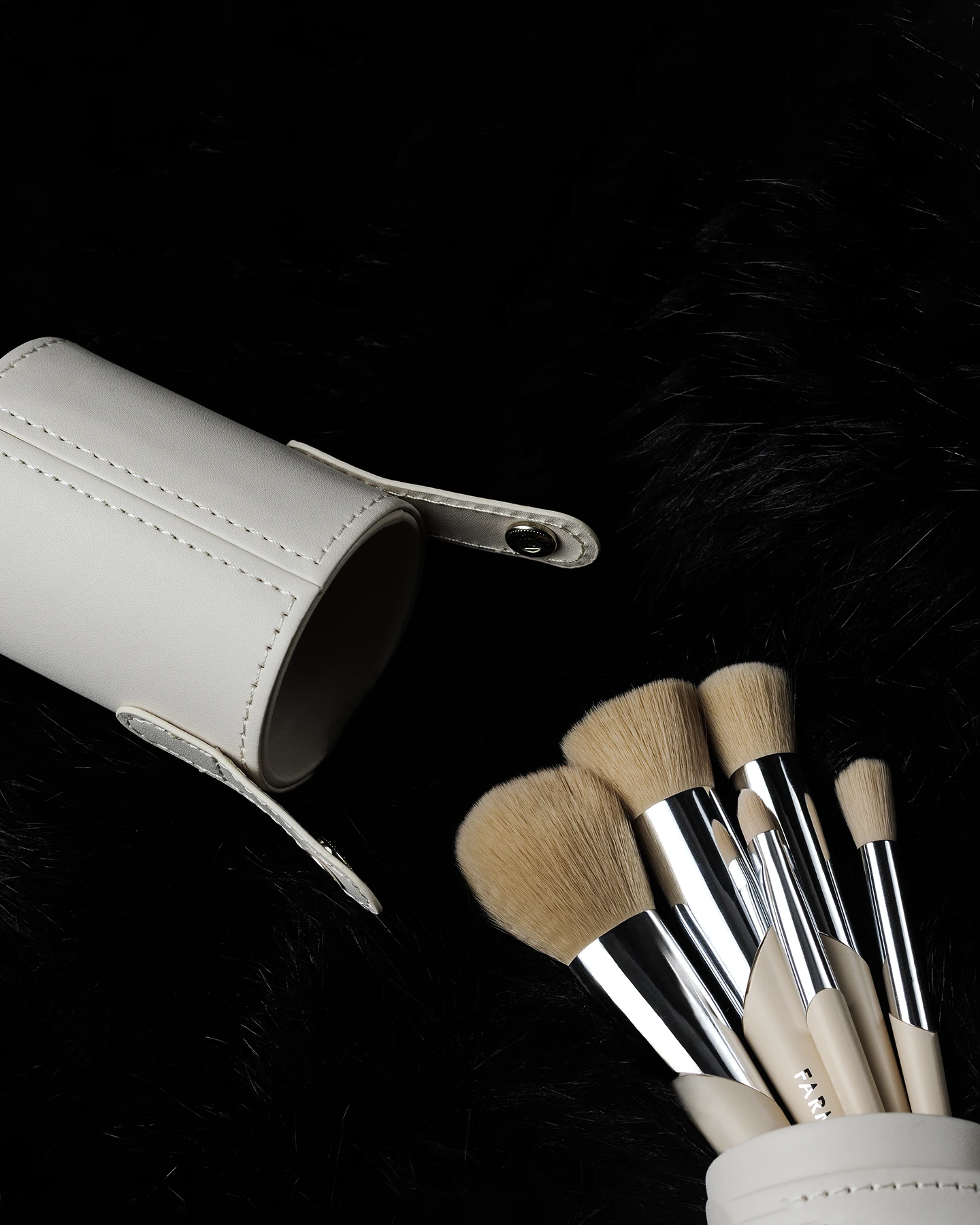 Fotografía cenital de set de brochas de maquillaje Farmasi con mangos de color beige y estuche cilíndrico de piel beige abierto, sobre fondo de piel sintética negra.