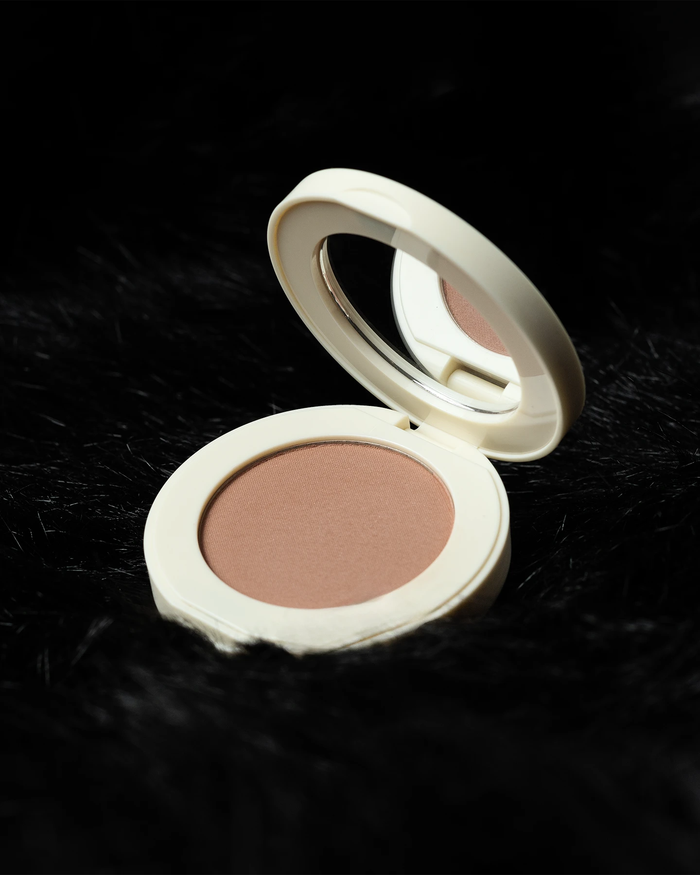 Toma cenital en ángulo de rubor compacto abierto de color coral rosado en estuche beige con espejo integrado, descansando sobre fondo de piel sintética negra texturizada.