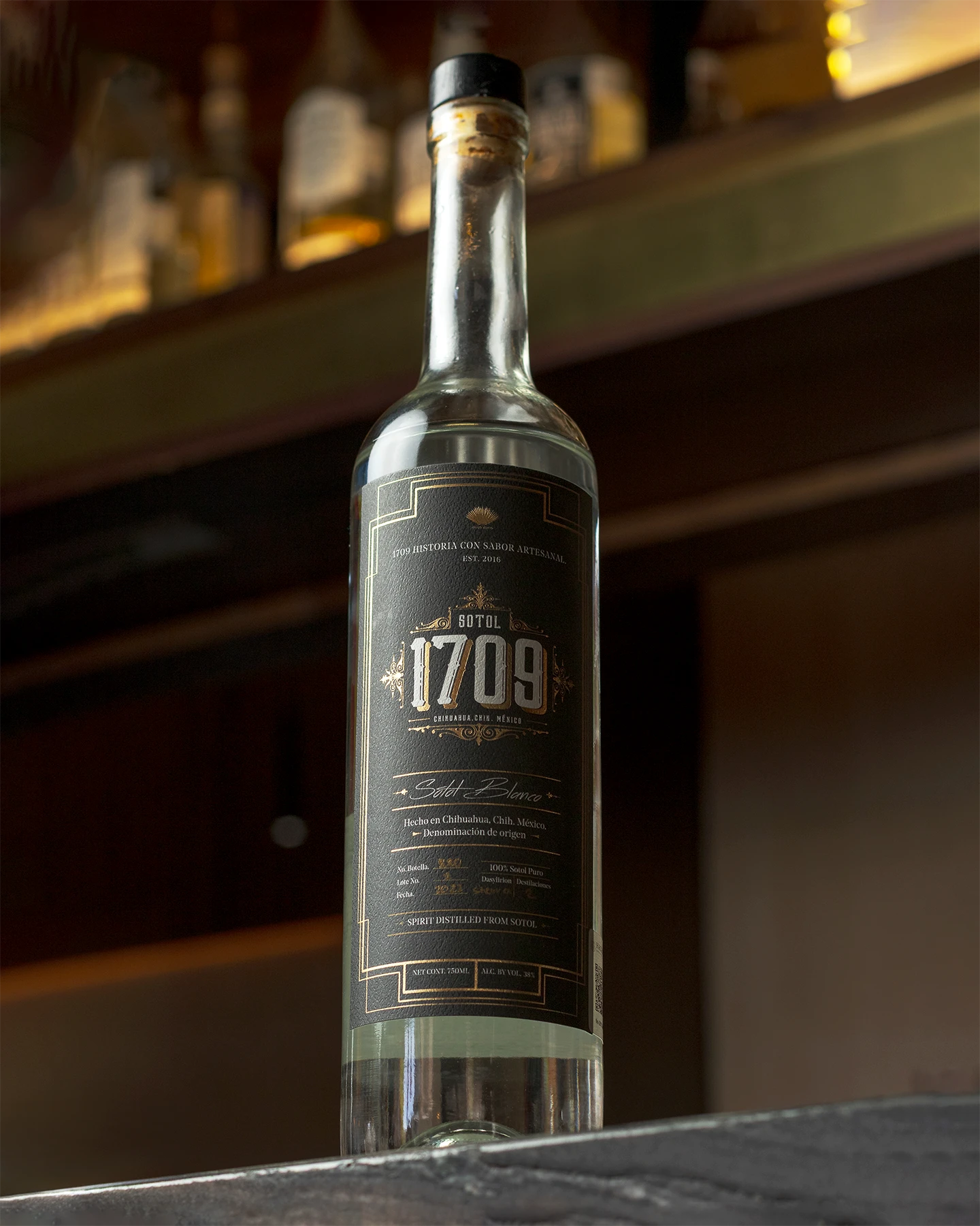 Fotografía editorial de una Botella de Sotol Blanco 1709 con etiqueta negra y dorado, en un ambiente de bar o barra desenfocado. Se lee 'Sotol Blanco' y 'Hecho en Chihuahua, Chih. México'.