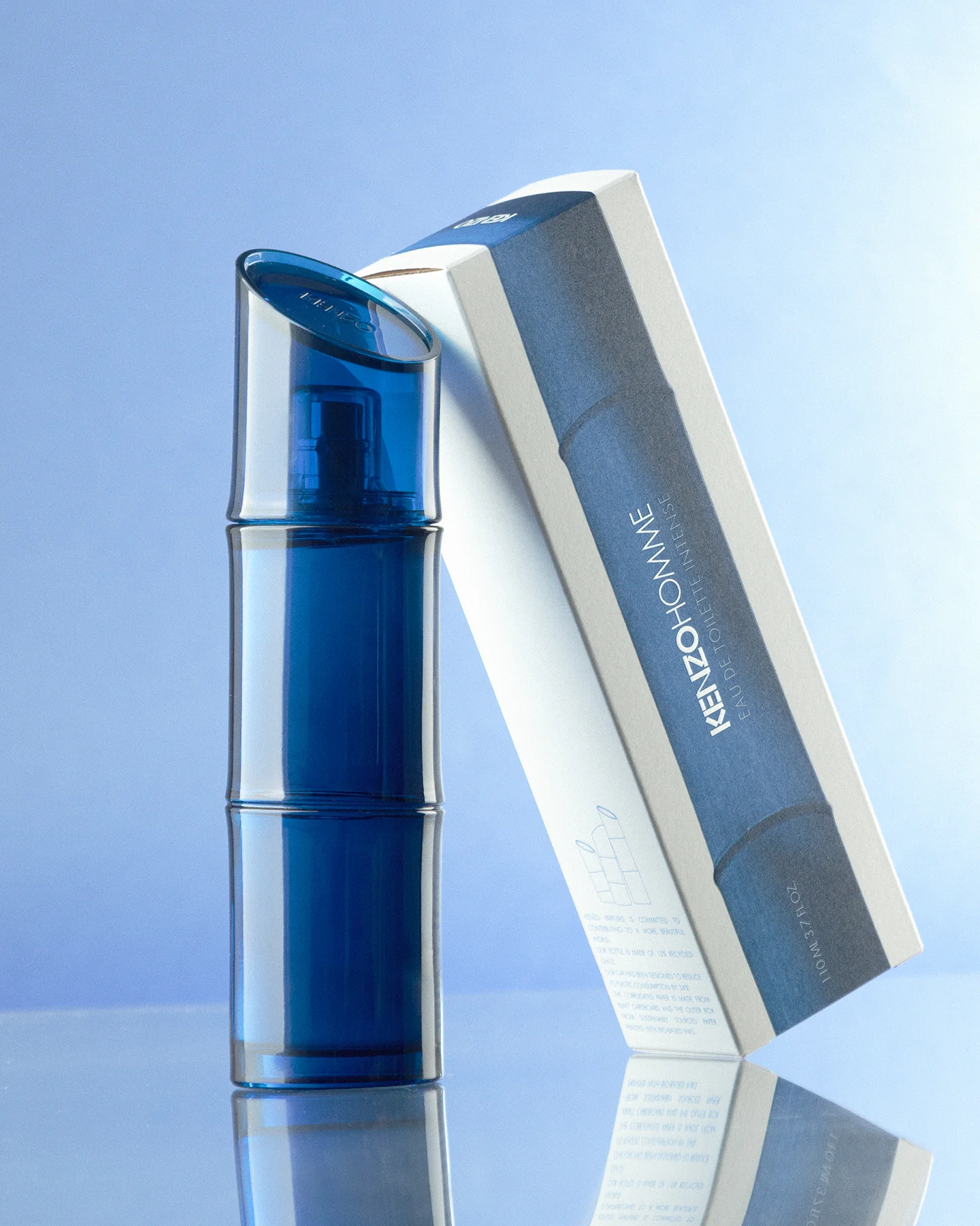 Fotografía de producto premium de botella de Kenzo Homme Eau de Toilette Intense con diseño de bambú azul y su caja blanca inclinada, sobre superficie reflectante con iluminación de estudio.