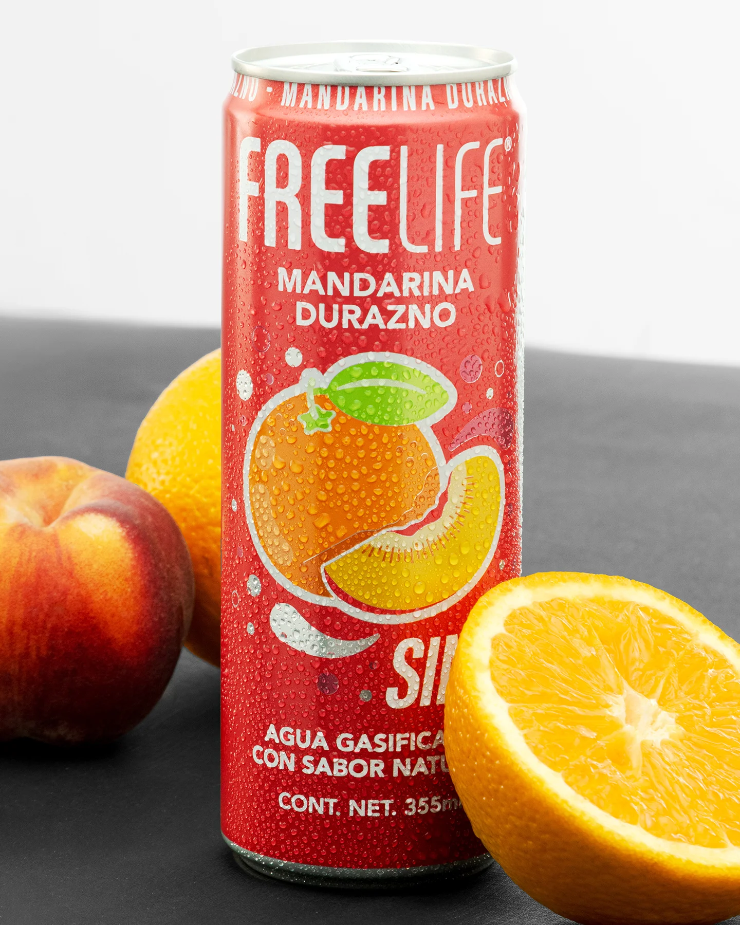 Fotografía de producto publicitaria de una lata helada y sudada Freelife Mandarina Durazno de 355ml, con rodaja de naranja fresca, naranja entera y durazno, sobre superficie negra.