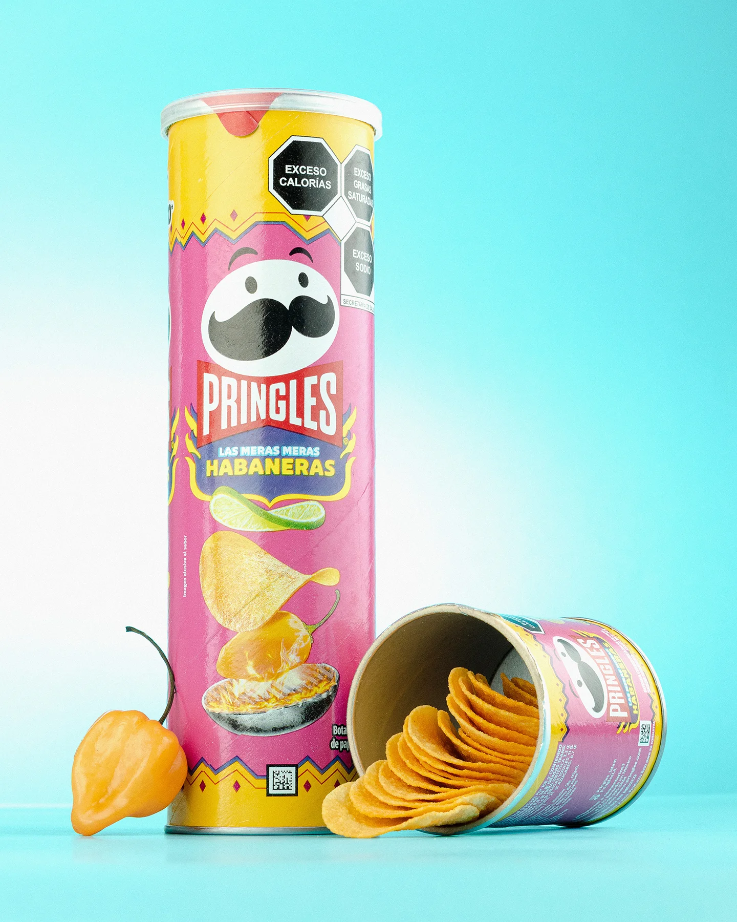 Fotografía publicitaria de lata de papas Pringles Las Meras Meras Habaneras junto a lata acostada con papas derramadas y un chile habanero fresco sobre fondo azul claro.