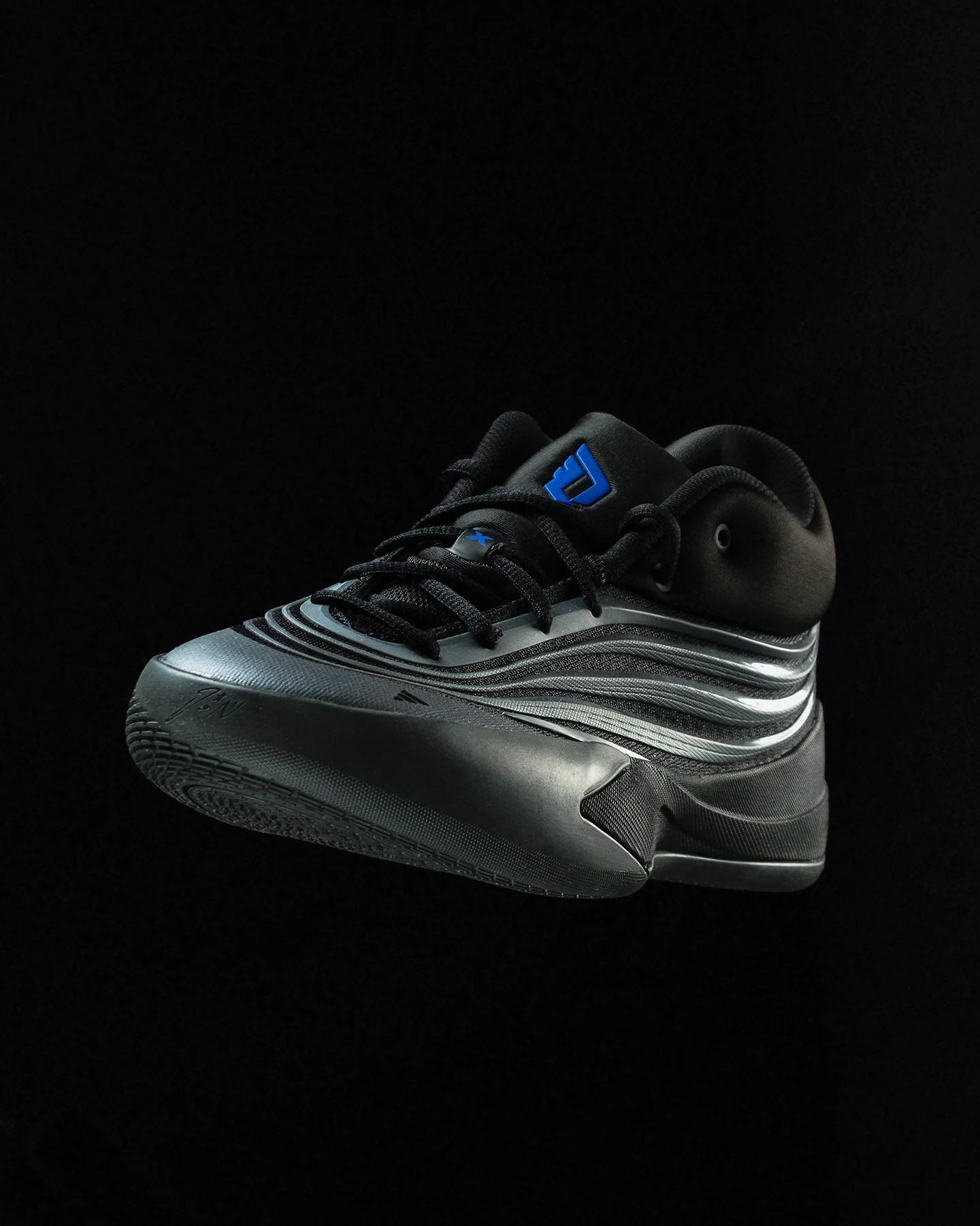 Toma frontal elevada de tenis Adidas negros y grises flotando sobre fondo negro, destacando el sistema de agujetas y el logo Adidas azul brillante en la lengüeta.