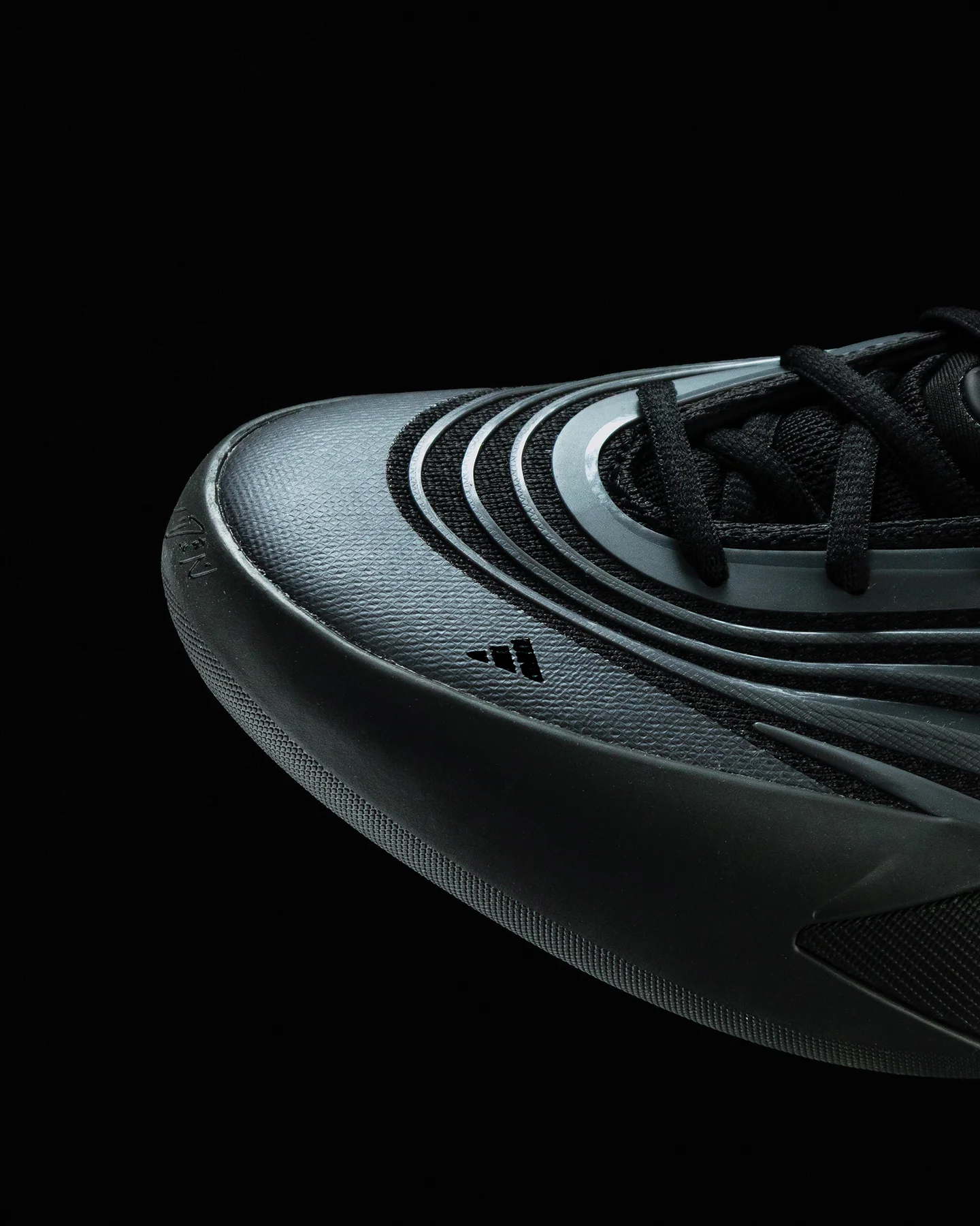 Detalle macro de la puntera texturizada de tenis Adidas negros con líneas de diseño futurista gris oscuro y pequeño logo de Adidas, con fondo negro.