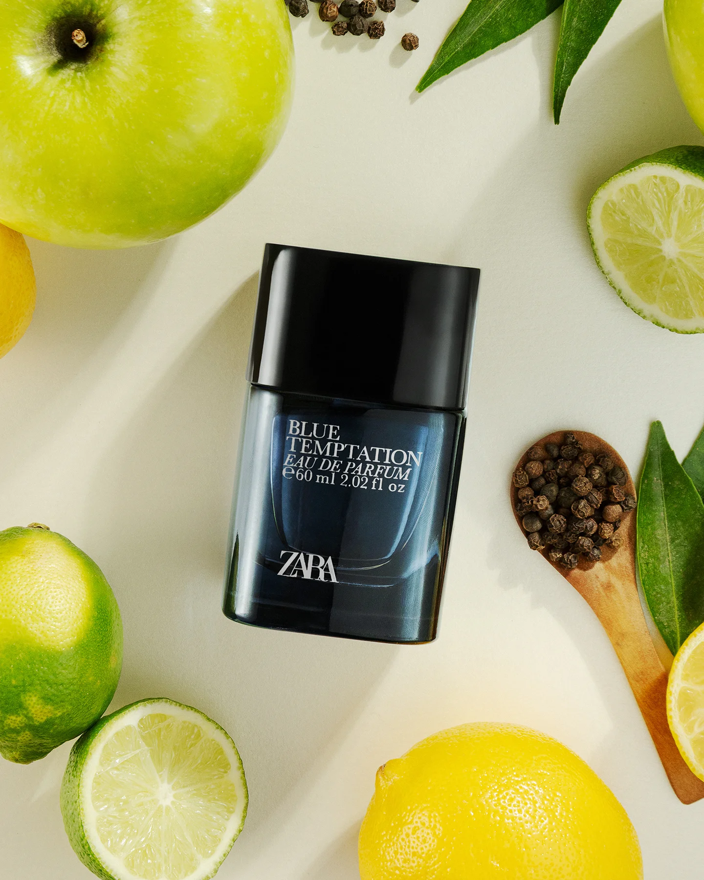 Fotografía cenital de perfume Zara Blue Temptation Eau de Parfum rodeado de manzanas verdes enteras, limones, rodajas de lima y una cuchara de madera con granos de pimienta negra sobre fondo claro.