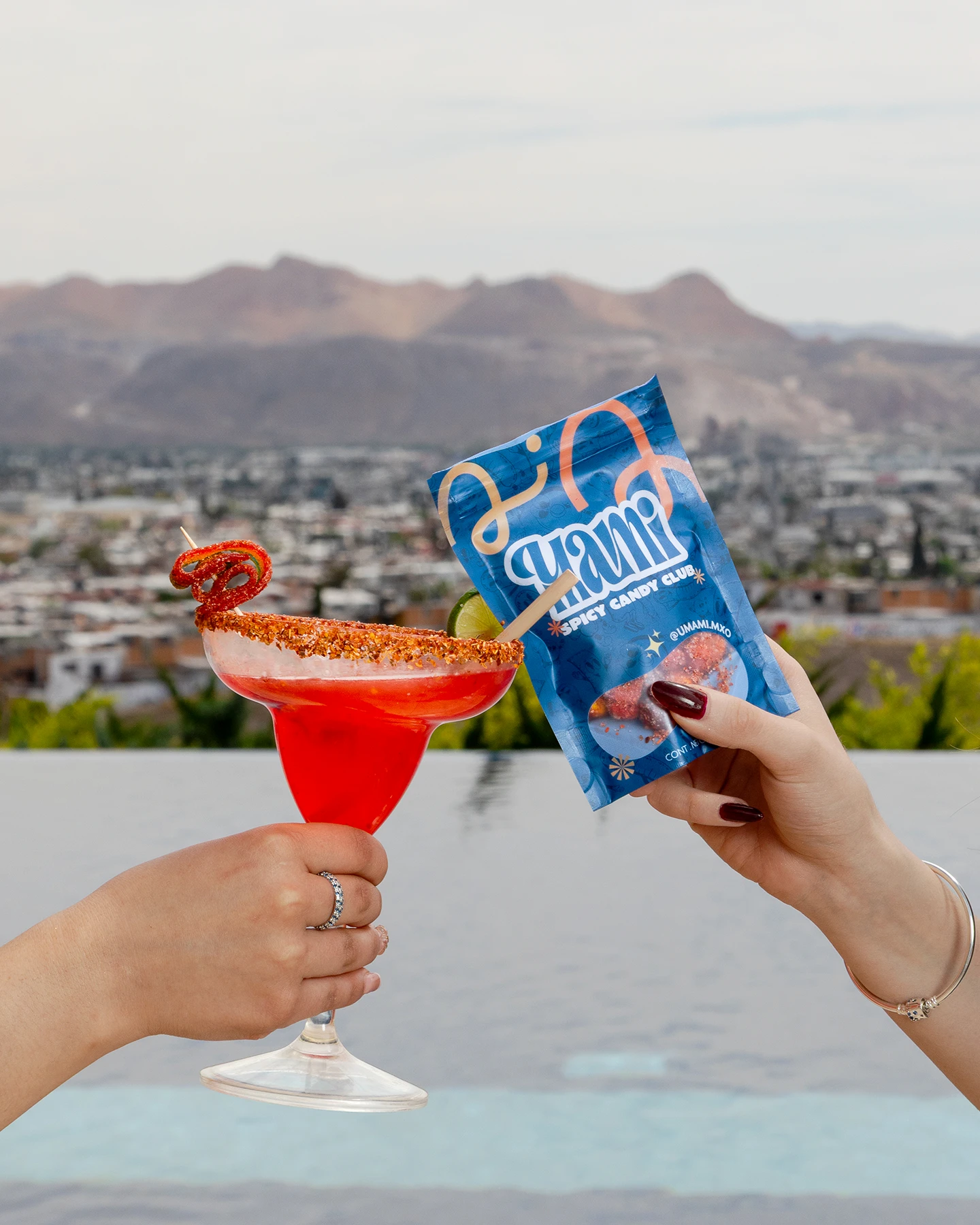 Brindis con margarita escarchada y bolsa de dulces picantes con paisaje urbano y montañas de Chihuahua al fondo.